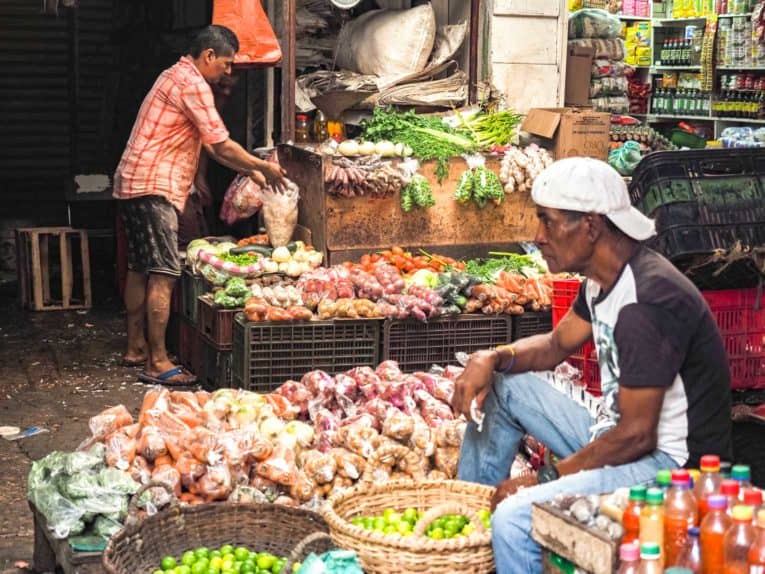Un homme est assis devant un stand de fruits et légumes.